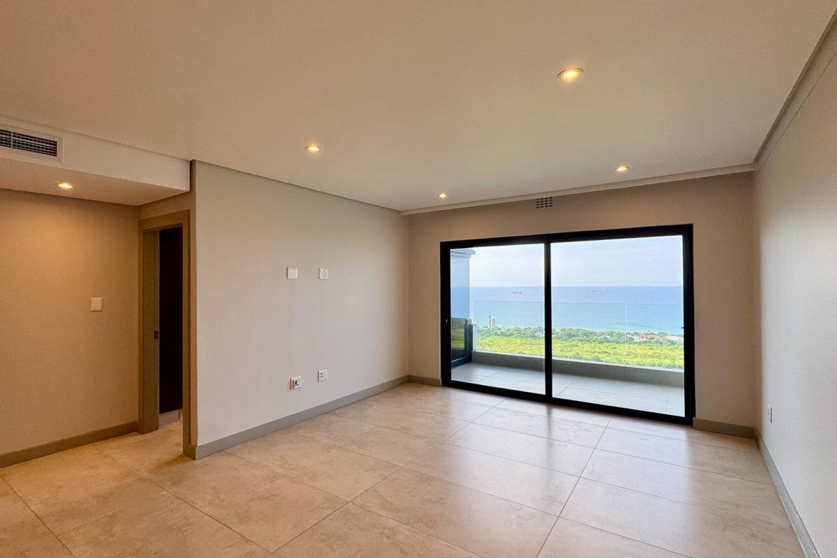 Spacious, modern living area featuring neutral tiled floors, recessed lighting, ducted air conditioning, and large sliding doors opening to a balcony with ocean views.