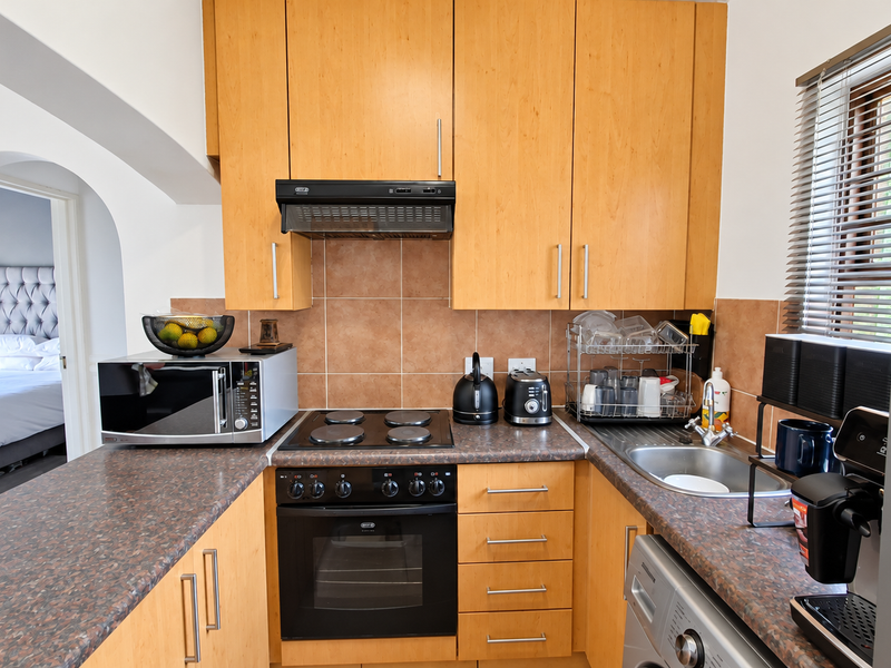A functional kitchen featuring light wood cabinetry, laminate countertops, a built-in oven with hob, and a tiled backsplash, with an adjacent view into the bedroom.