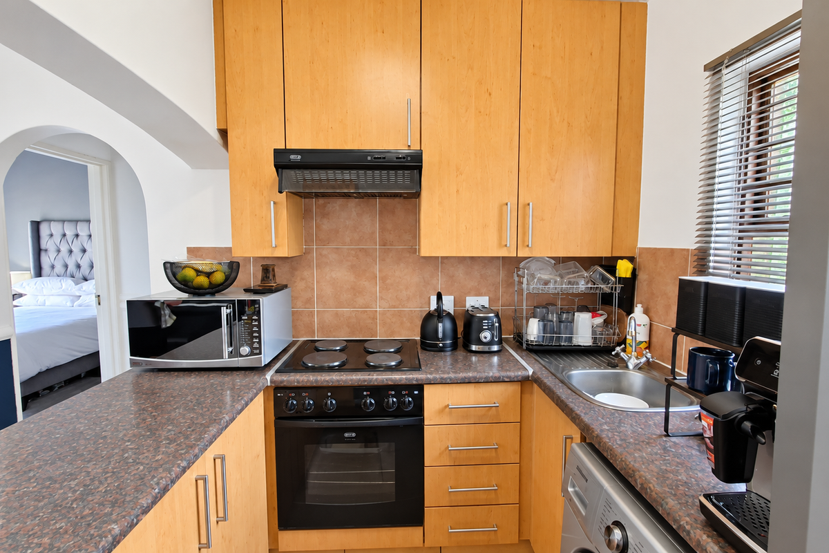 A functional kitchen featuring light wood cabinetry, laminate countertops, a built-in oven with hob, and a tiled backsplash, with an adjacent view into the bedroom.