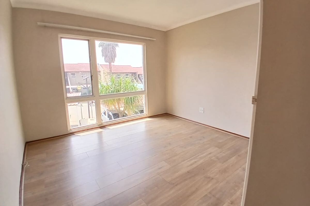 A bright, empty bedroom featuring light wood-look laminate flooring, neutral walls, and a large window providing natural light and a view of the complex.