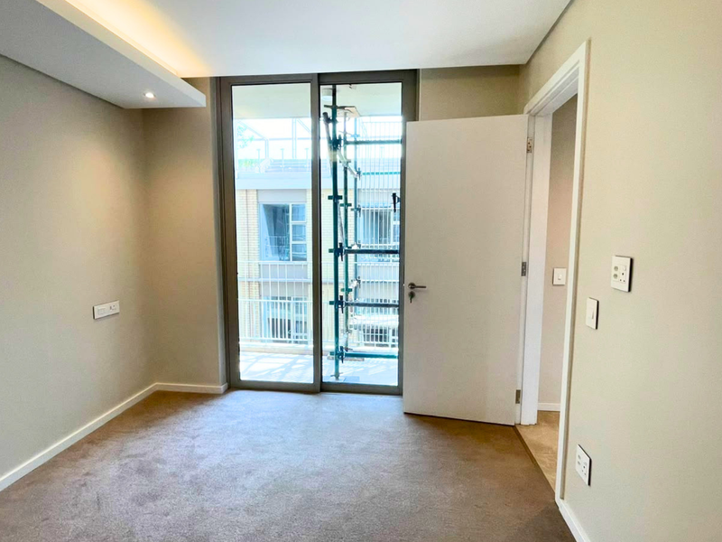 A bright, empty bedroom featuring neutral walls, carpeted flooring, recessed ceiling lighting, and a sliding glass door providing access to a balcony.