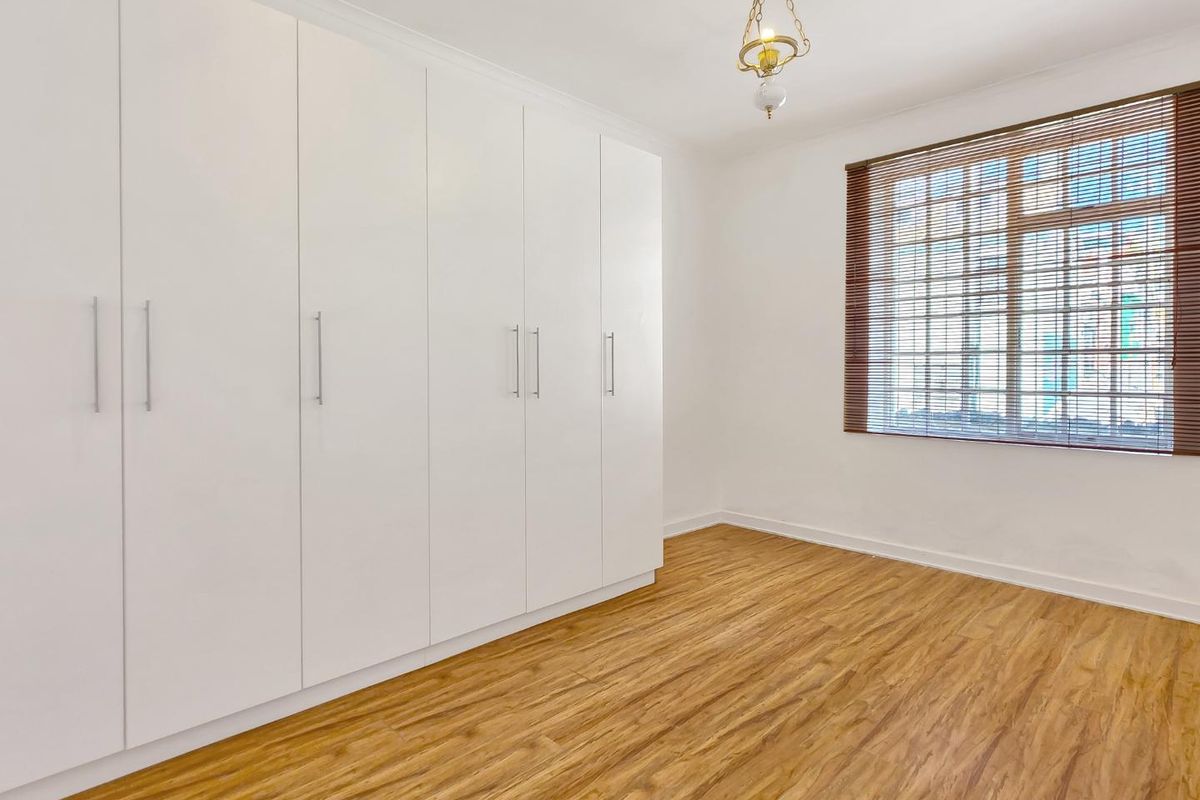 A bright, empty bedroom featuring light timber laminate flooring, extensive white built-in wardrobes, and window blinds.