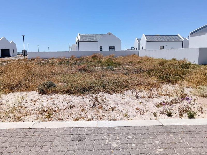 A vacant residential plot featuring sandy terrain and a boundary wall, situated in a developed neighborhood with paved street access.