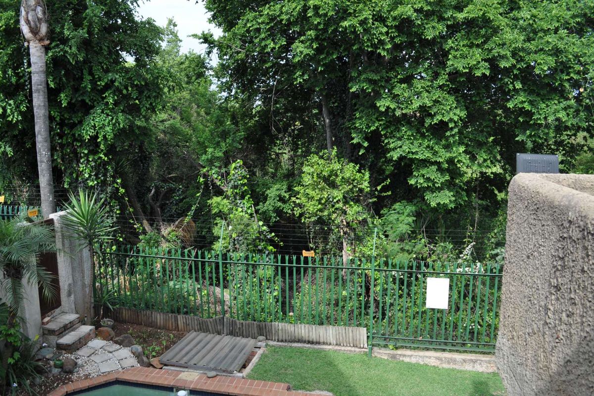 Rear garden area featuring a swimming pool, brick-edged paving, and metal security fencing bordered by dense mature trees.