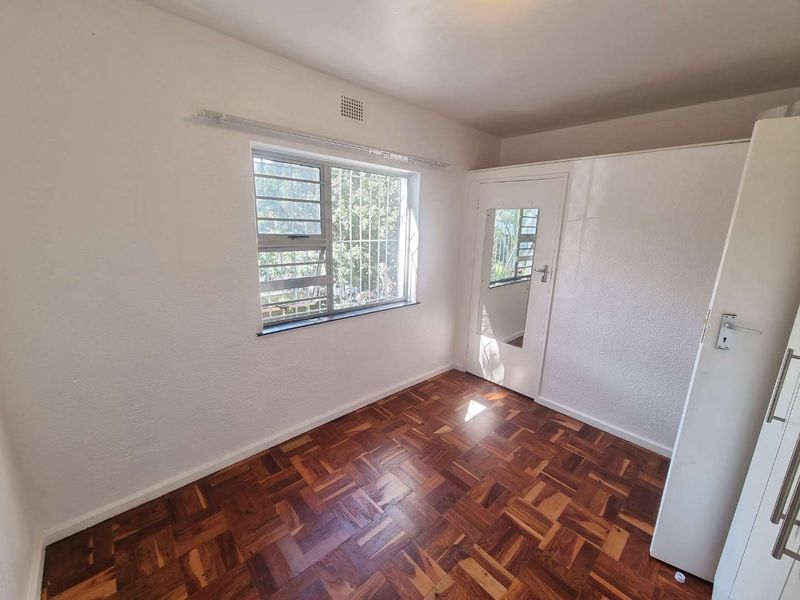 A bright, empty bedroom featuring classic parquet flooring, a built-in cupboard with a mirror, and a window fitted with security bars.