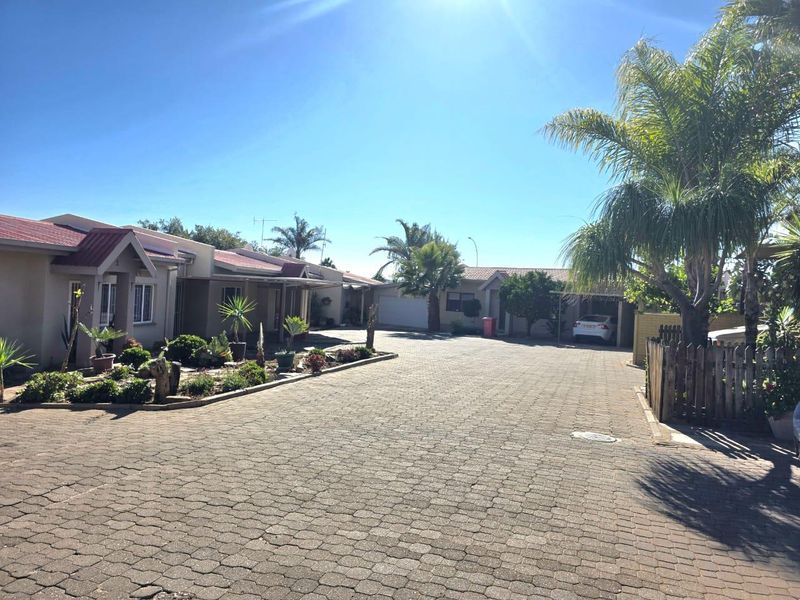 A view of the townhouse complex entrance featuring a paved driveway, established garden beds, and multiple garage access points under a clear sky.
