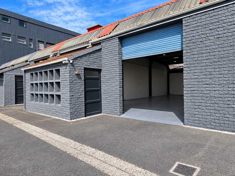 Exterior view of an industrial warehouse facility featuring grey brick facade, multiple roller shutter doors, and paved driveway access.