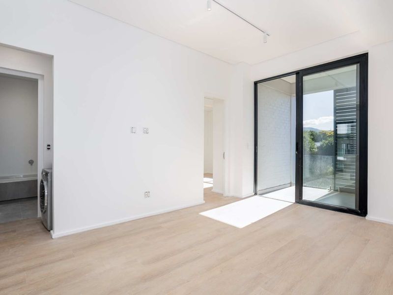 A bright, empty living area featuring modern laminate flooring, track lighting, and a large sliding glass door leading to an outdoor balcony.