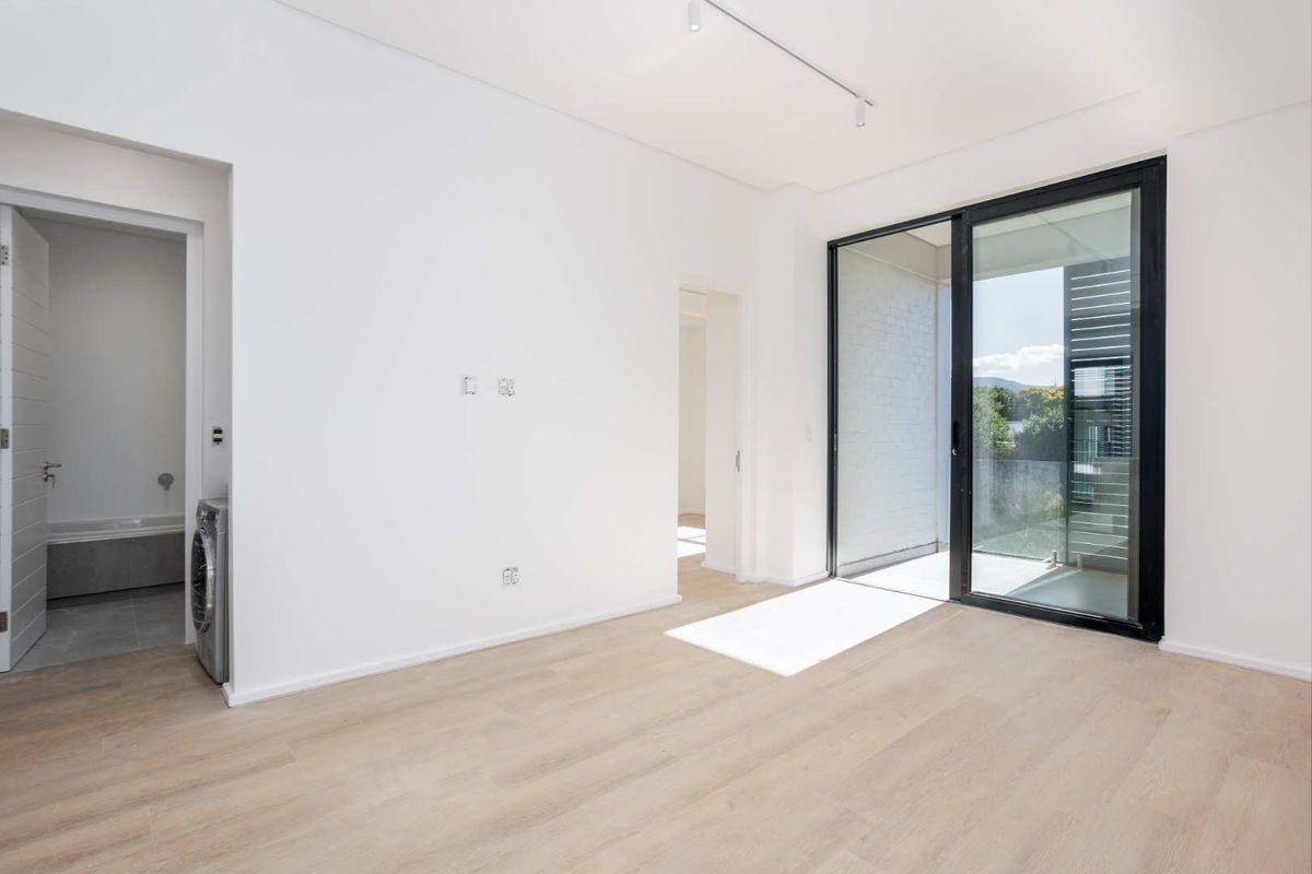 A bright, empty living area featuring modern laminate flooring, track lighting, and a large sliding glass door leading to an outdoor balcony.