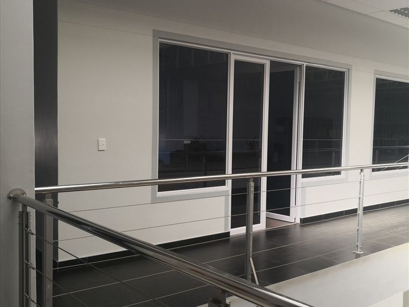 A modern industrial hallway featuring a stainless steel balustrade, dark tiled flooring, and clean white walls with aluminium framed windows and doors.