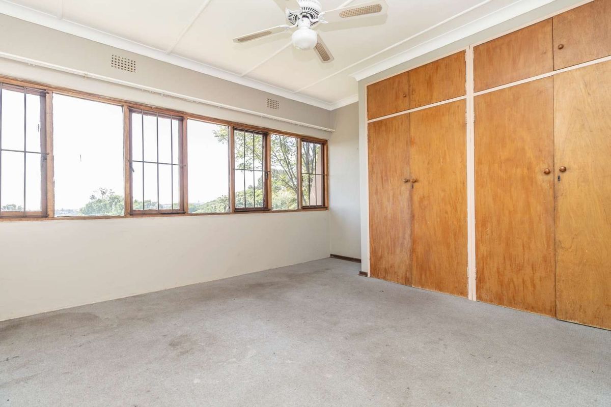 A spacious, empty bedroom featuring built-in wooden cupboards, a ceiling fan, and large windows with security bars, offering a blank canvas for renovation.