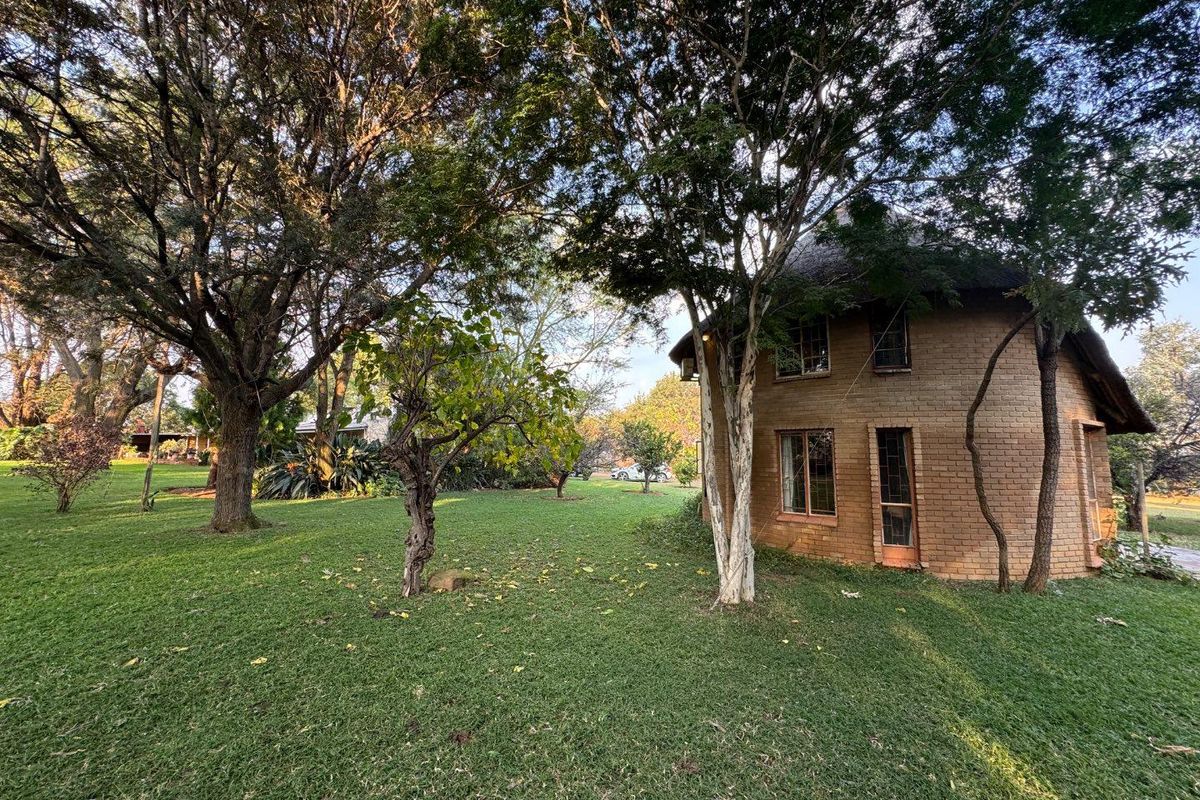 A view of the exterior rear of the property, featuring a brick structure with a thatched roof set within a spacious, tree-lined garden.