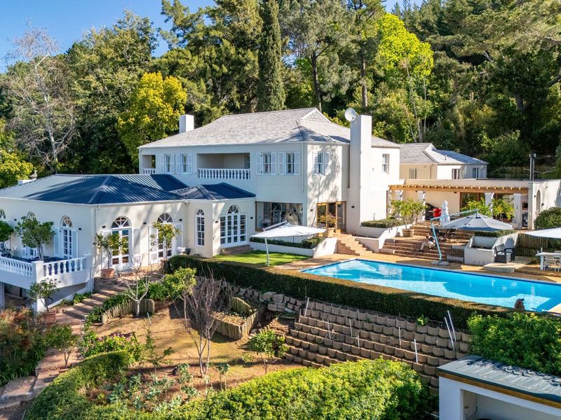 Aerial view of a large multi-level residence featuring a swimming pool, terraced gardens, retaining walls, and a spacious balcony set against a wooded backdrop.