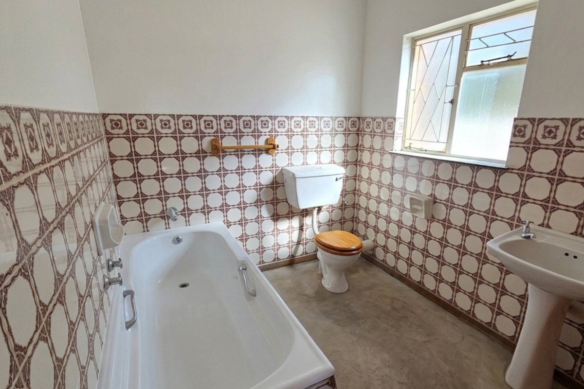 A dated bathroom featuring a standard bathtub, pedestal basin, and toilet, finished with vintage patterned wall tiles and a simple window.