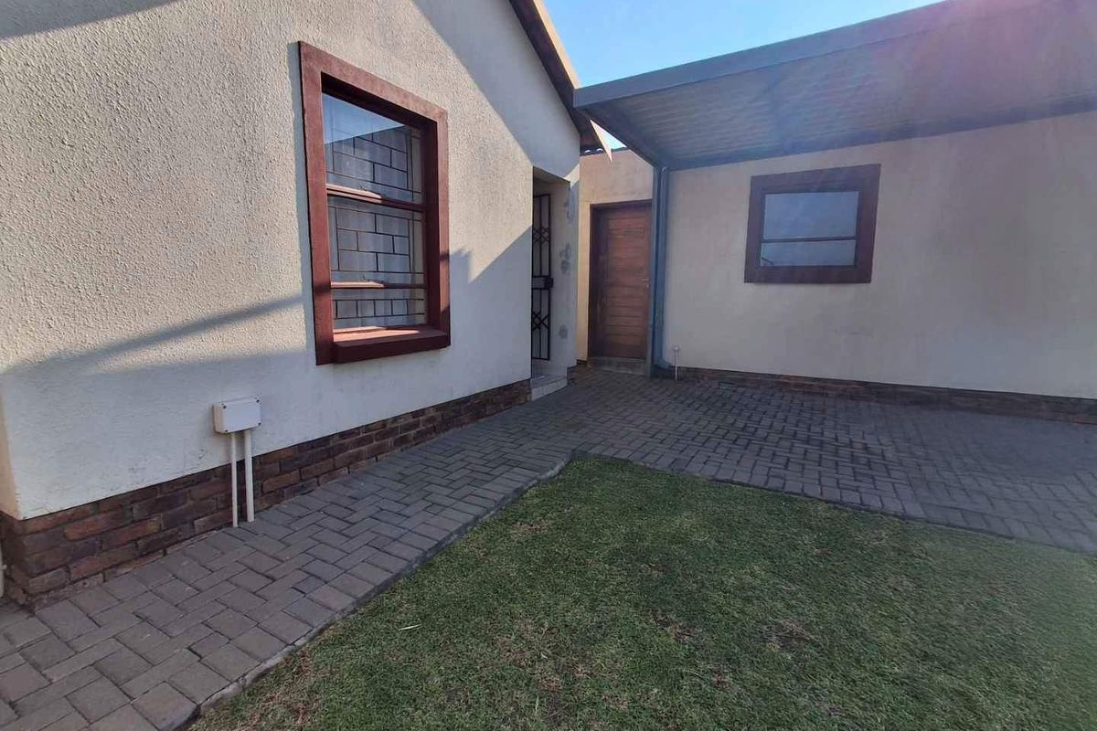 A paved rear courtyard area featuring a covered patio section, brick-trimmed exterior walls, and a small lawn.