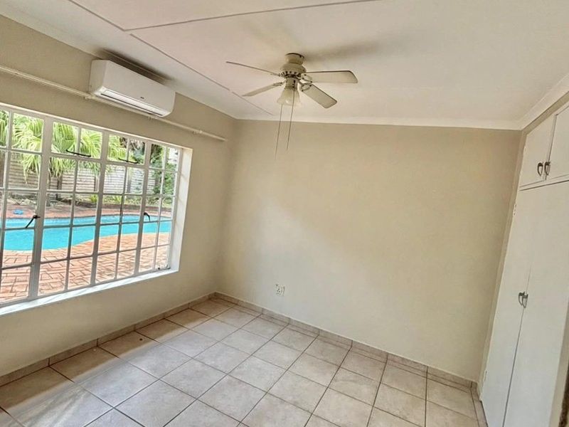 An empty bedroom featuring tiled flooring, built-in cupboards, a ceiling fan, and a split-system air conditioner, with a view overlooking the pool area.