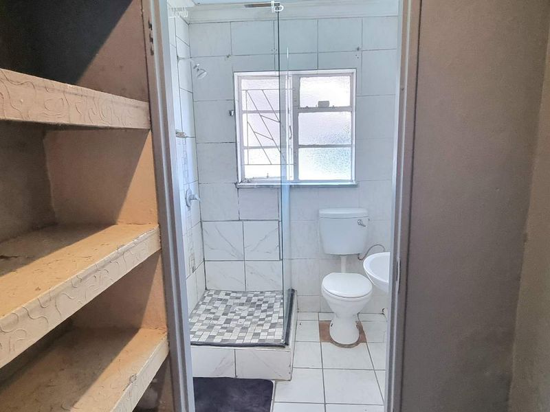 A functional bathroom featuring a shower enclosure, toilet, and pedestal basin, adjacent to a storage area with built-in shelving.