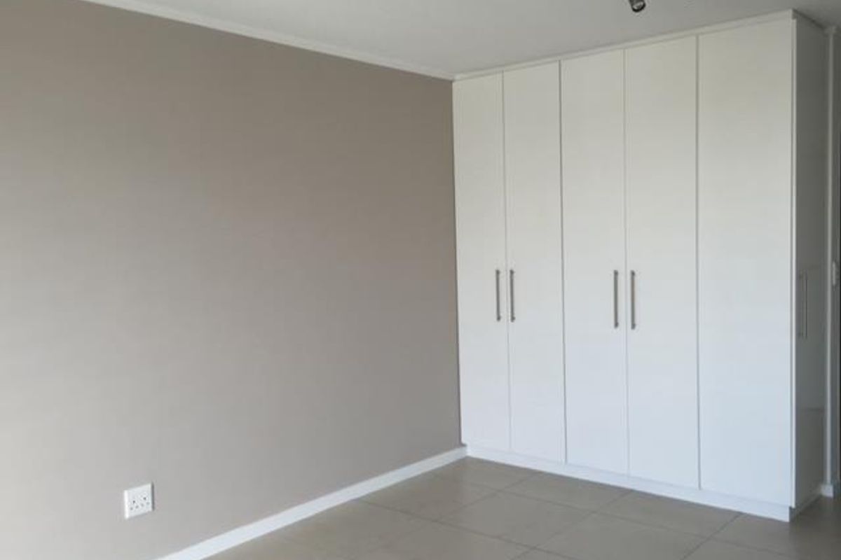 A clean, empty bedroom featuring modern white built-in cupboards, neutral wall tones, and tiled flooring with contemporary track lighting.
