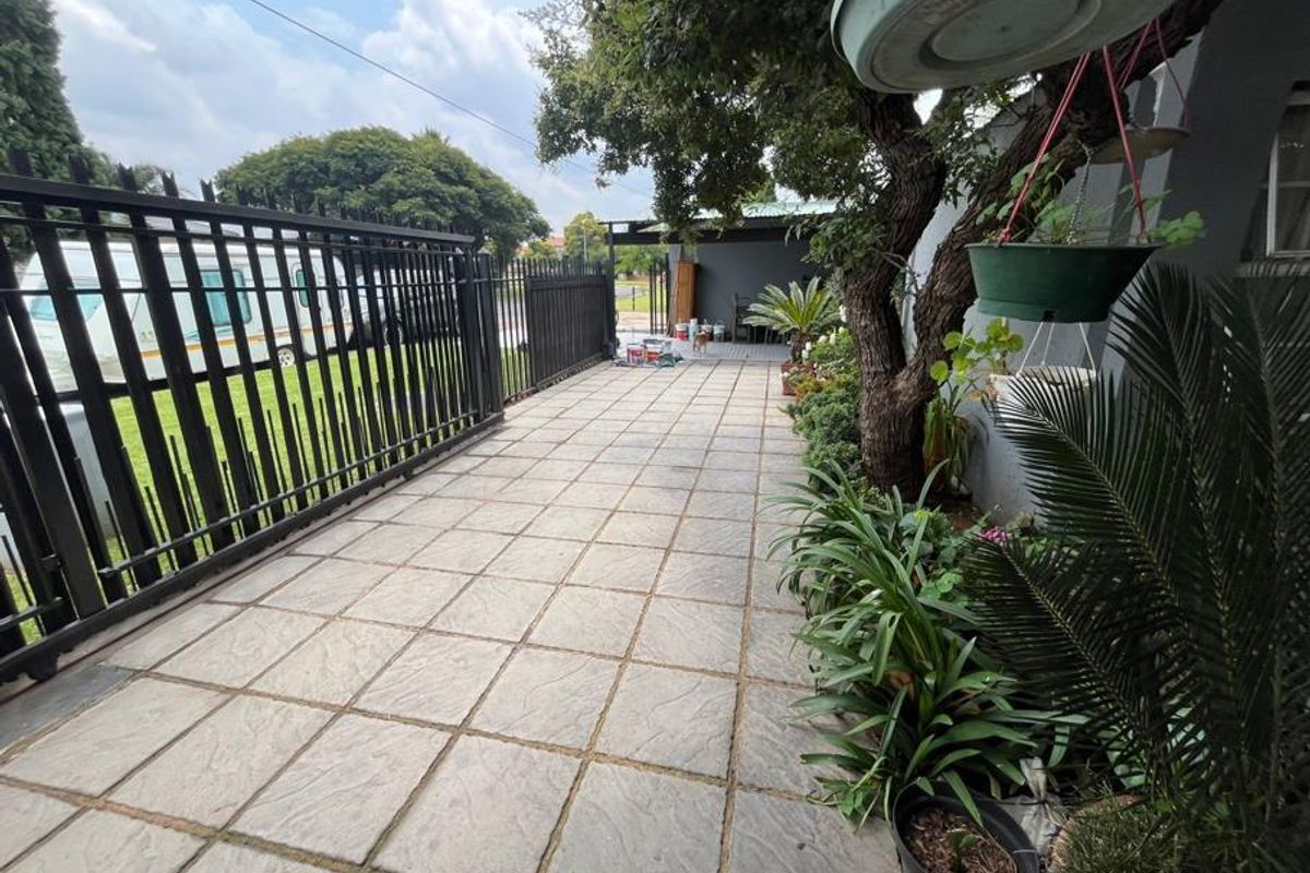 A paved side walkway featuring a metal security fence, mature trees, and established garden beds leading toward a covered outdoor area.