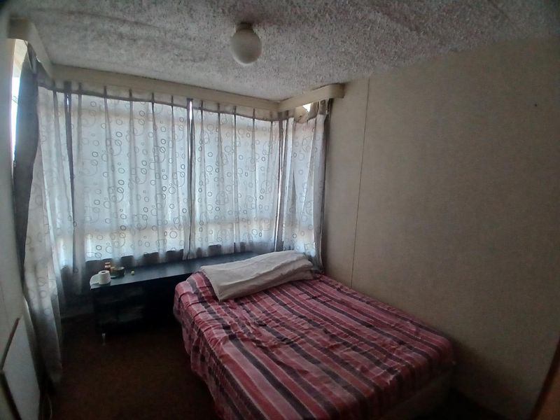 A compact bedroom featuring a large window with curtains and a textured ceiling, requiring cosmetic updates to improve its overall condition and appeal.