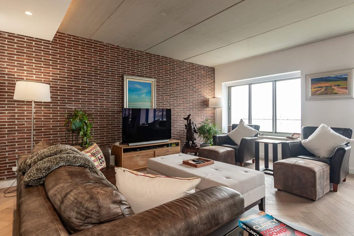 A spacious living room featuring an exposed brick feature wall, herringbone timber flooring, and large windows providing ample natural light.