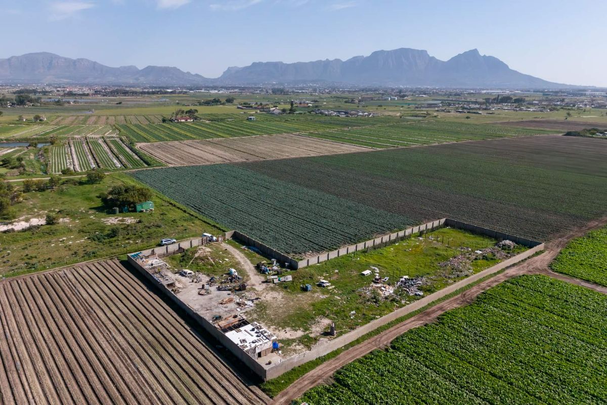 Aerial view of a vacant residential plot enclosed by a perimeter wall, situated within a rural agricultural landscape with mountain views.
