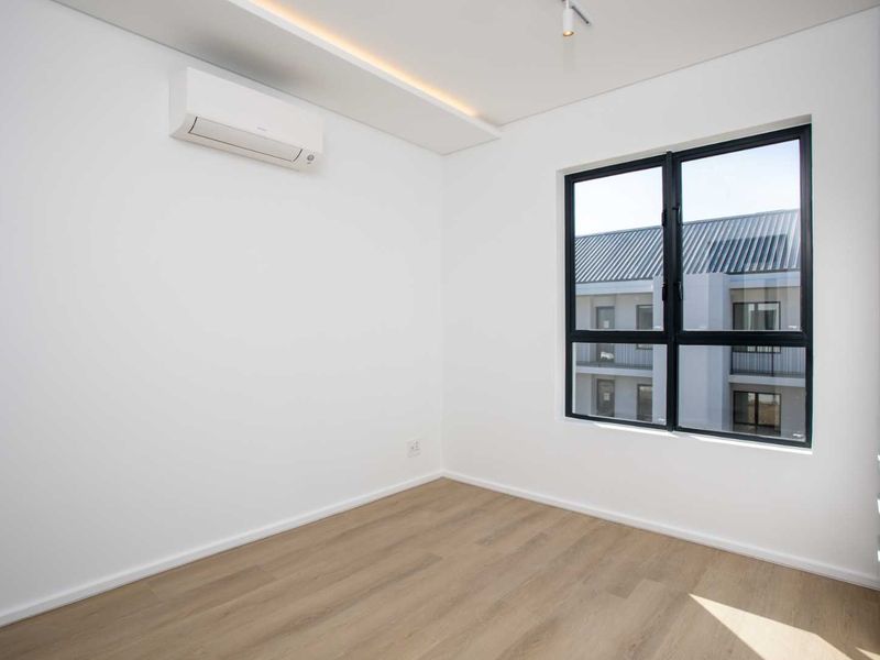 A bright, modern, and empty bedroom featuring light wood-look laminate flooring, a split-system air conditioner, and a large black-framed window.