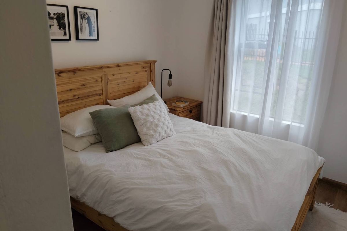 A cozy bedroom featuring neutral walls, wooden flooring, and a window with sheer curtains, providing a bright and comfortable living space.