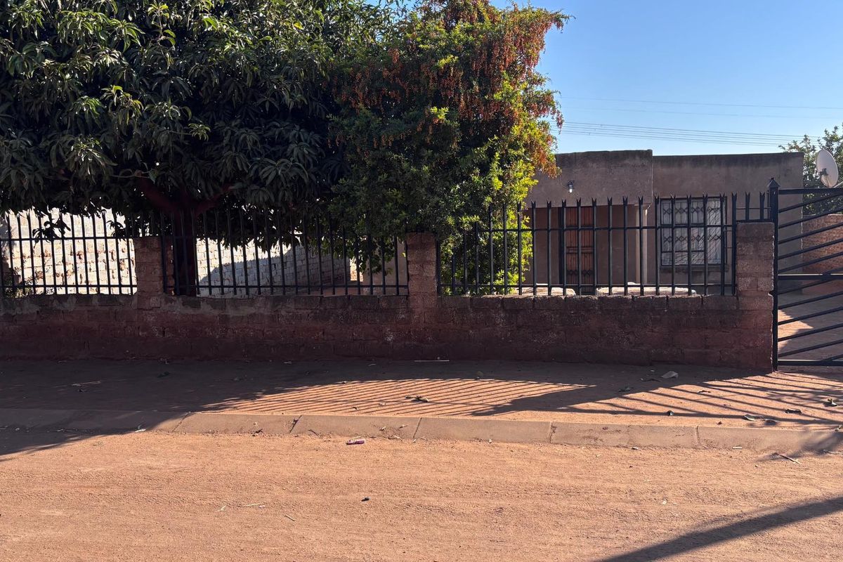 Front view of the property featuring a brick boundary wall, metal security fencing, and a gated entrance with mature trees providing shade.