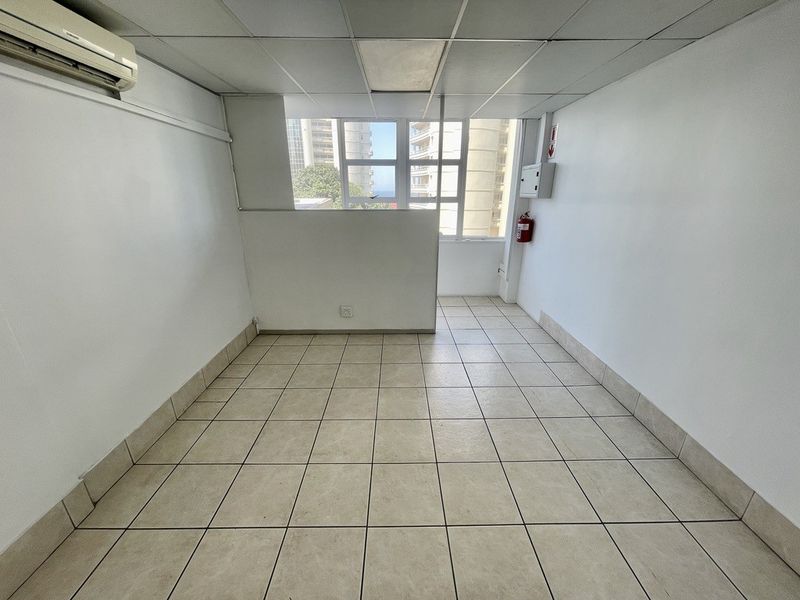A clean, empty office space featuring ceramic floor tiles, a suspended ceiling, and a wall-mounted air conditioning unit with natural light.