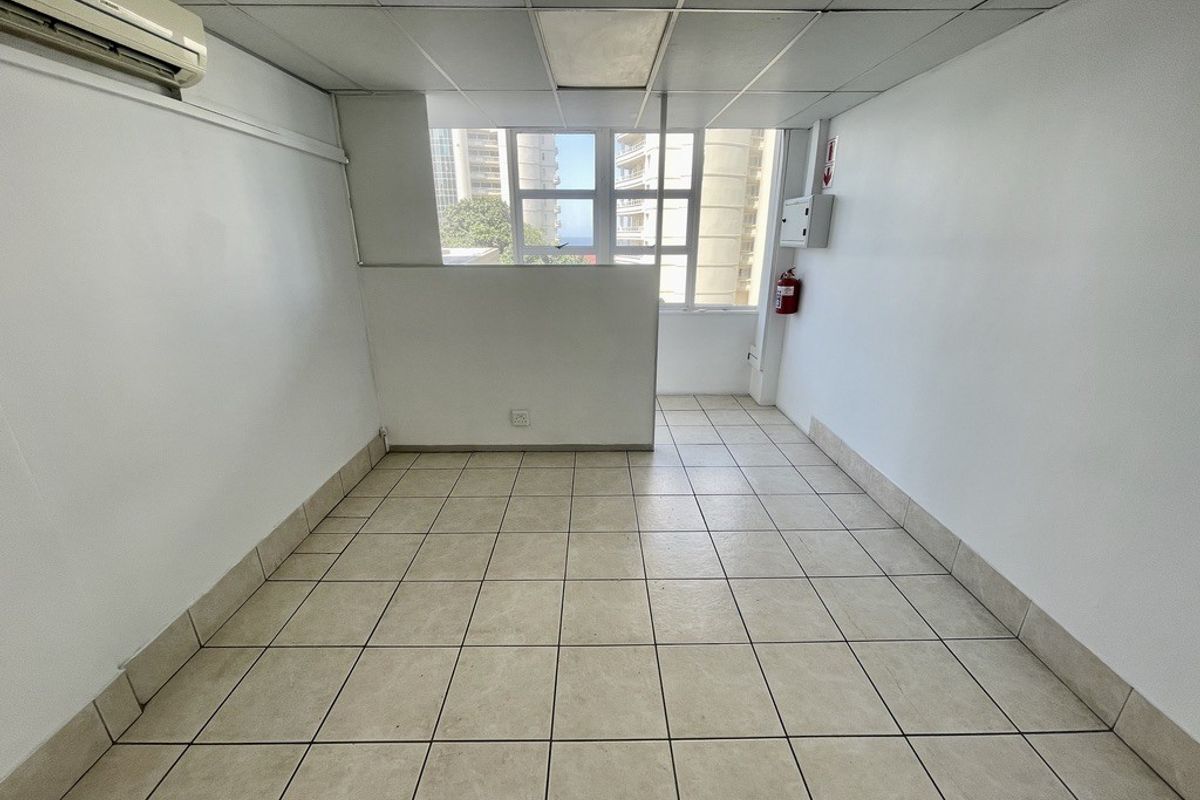 A clean, empty office space featuring ceramic floor tiles, a suspended ceiling, and a wall-mounted air conditioning unit with natural light.