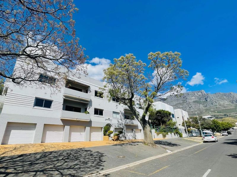 Front elevation of a multi-story apartment building featuring ground-level garages, private balconies, and a paved driveway, set against a mountain backdrop.