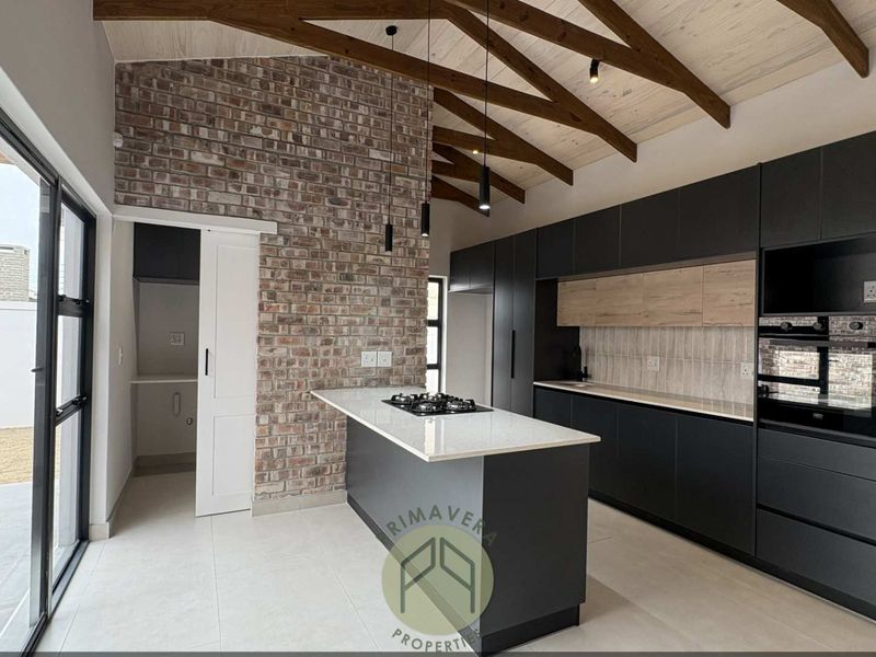 Modern kitchen featuring a central island with gas hob, exposed brick wall, timber vaulted ceiling, and sleek cabinetry, with an adjacent scullery area.