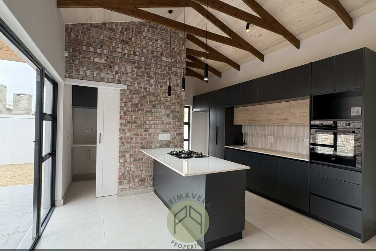 Modern kitchen featuring a central island with gas hob, exposed brick wall, timber vaulted ceiling, and sleek cabinetry, with an adjacent scullery area.