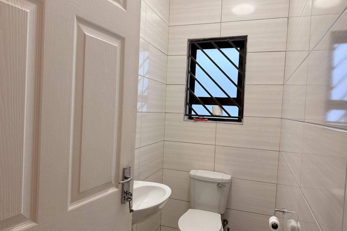 A compact, modern bathroom featuring neutral wall tiles, a wall-mounted basin, and a toilet with a window fitted with security bars.