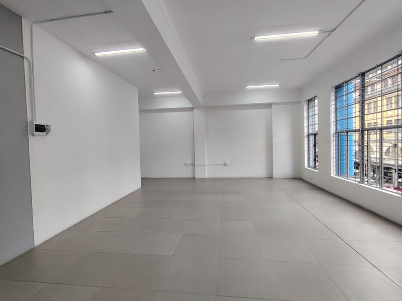 A bright, open-plan office space featuring neutral tiled flooring, large windows with security bars, and modern overhead lighting.