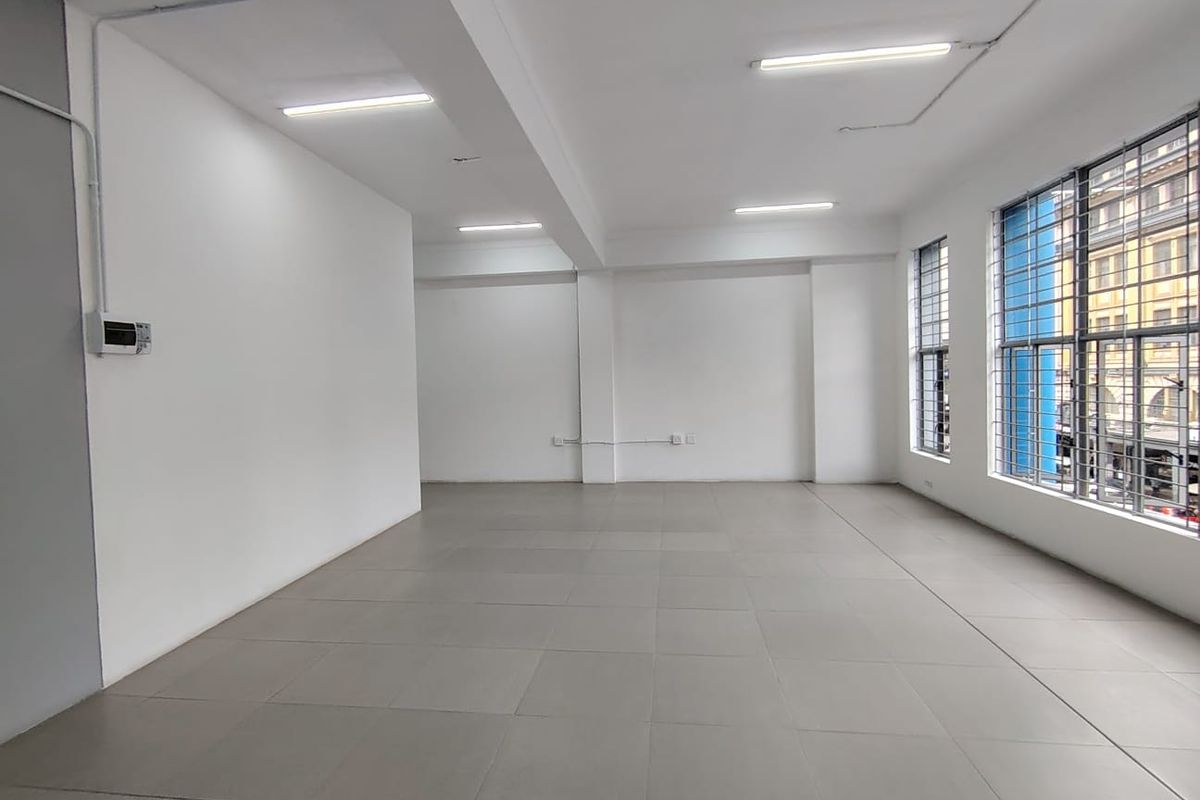 A bright, open-plan office space featuring neutral tiled flooring, large windows with security bars, and modern overhead lighting.