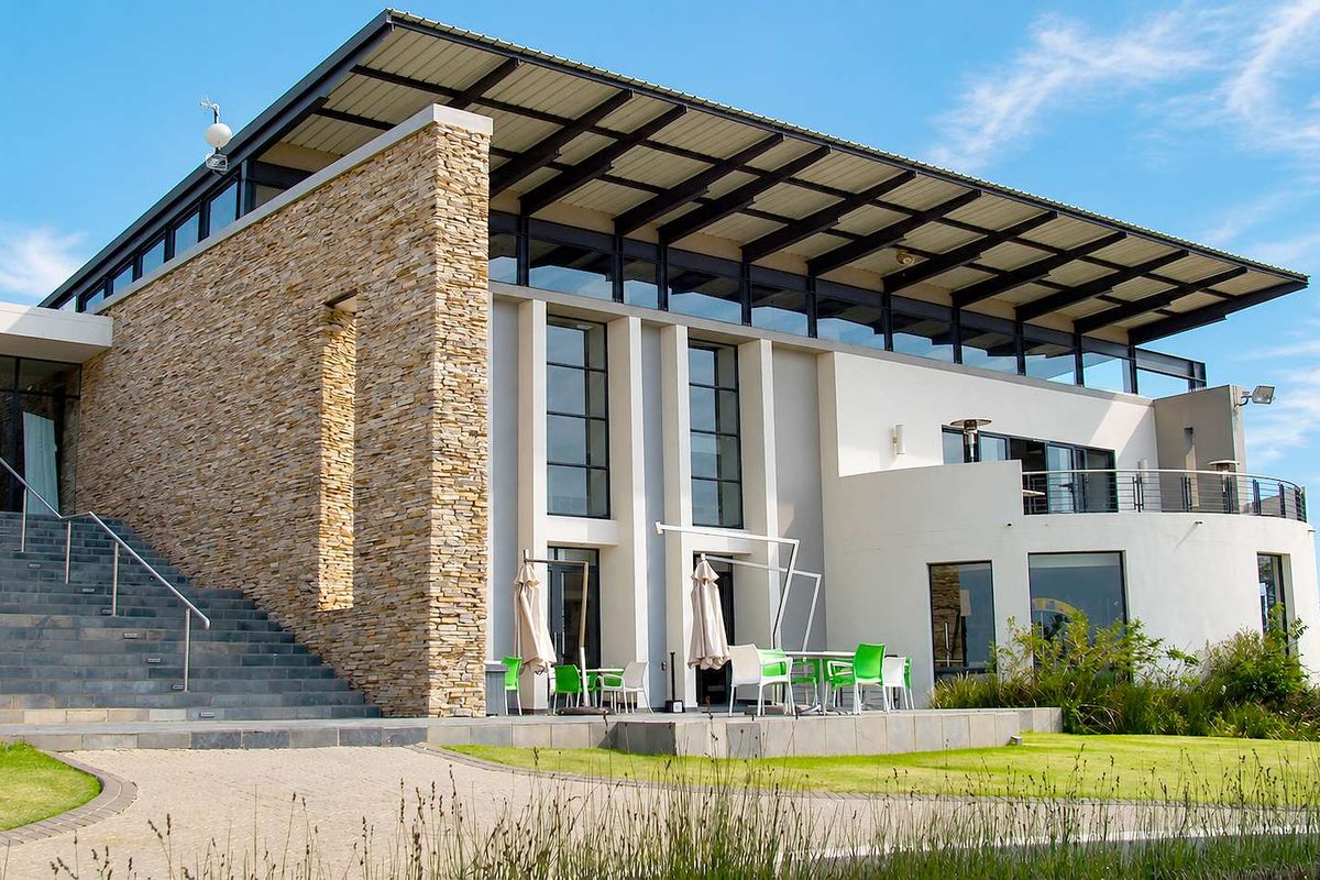 Modern architectural exterior featuring stone cladding, expansive glass windows, a large metal roof, and a spacious paved patio area.
