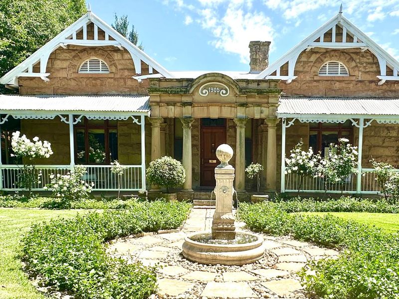 A charming historic stone house featuring a classic veranda, symmetrical gabled roof, and a beautifully landscaped front garden with a central stone fountain.