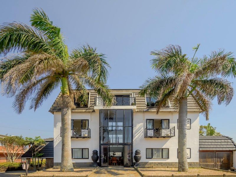 A striking multi-story residential facade featuring a prominent central glass atrium, wrought iron balconies, and mature palm trees framing the entrance.