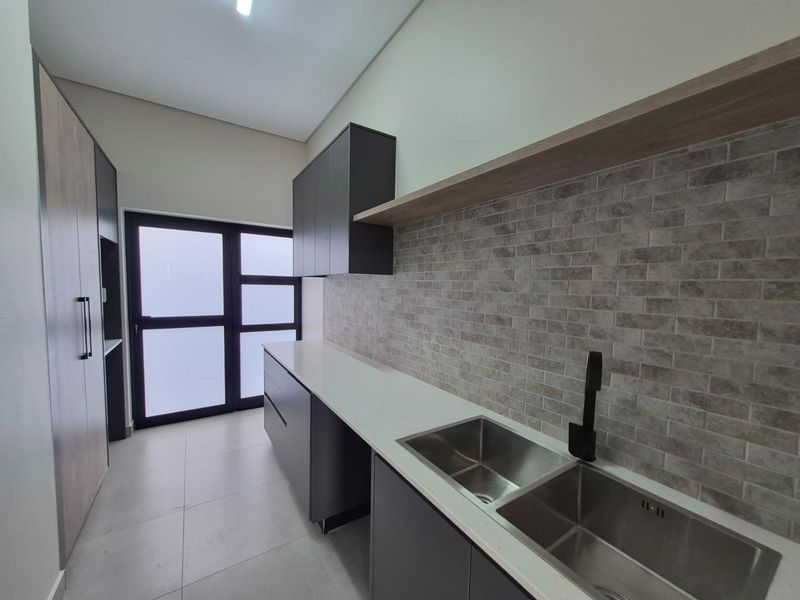 A modern, clean scullery featuring double stainless steel sinks, matte black fixtures, stone countertops, and ample built-in cabinetry with a stylish brick-tiled backsplash.