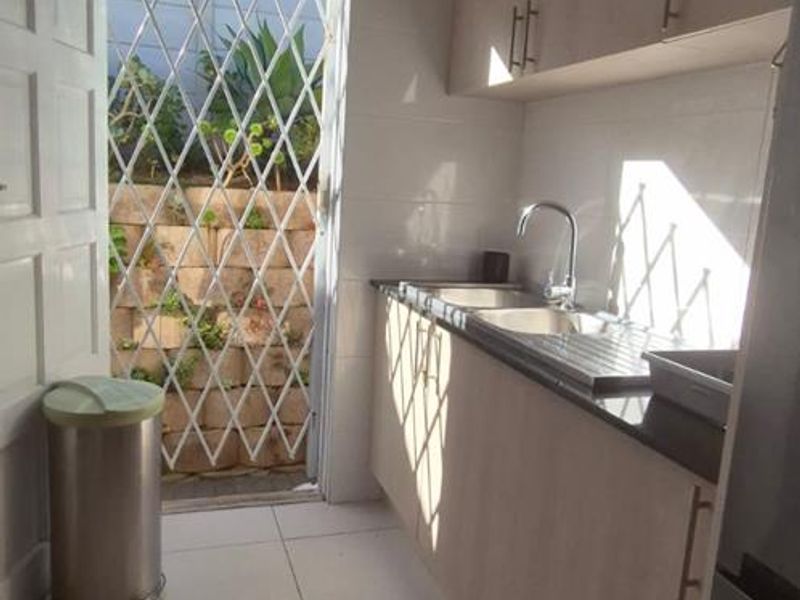 A functional scullery area featuring light wood-finish cabinetry, a stainless steel sink, and direct access to an outdoor space through a security gate.
