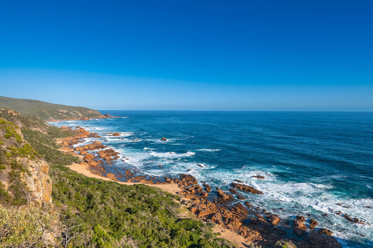 A scenic coastal view featuring rugged cliffs, rocky shoreline, and expansive ocean waters under a clear blue sky.