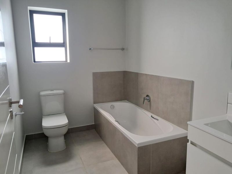 A clean, modern bathroom featuring a built-in bathtub, a white toilet, and a sleek vanity cabinet with neutral wall and floor tiling.