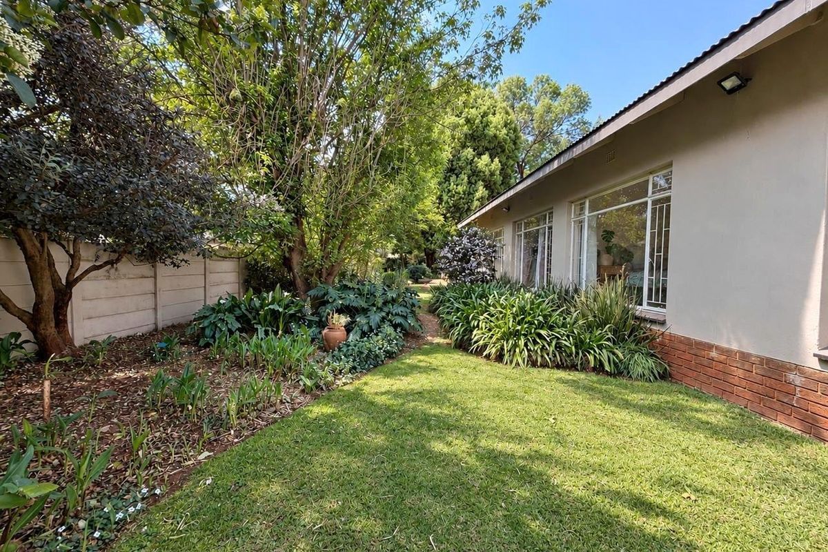 A well-maintained rear garden featuring a lush lawn, mature trees, established garden beds, and a brick-accented exterior wall with large windows.