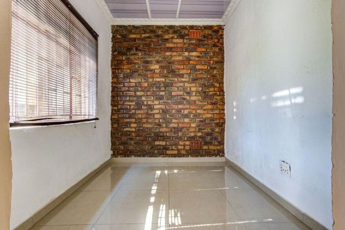 An empty bedroom featuring a rustic exposed brick feature wall, neutral tiled flooring, and venetian blinds, offering a blank canvas for personalization.