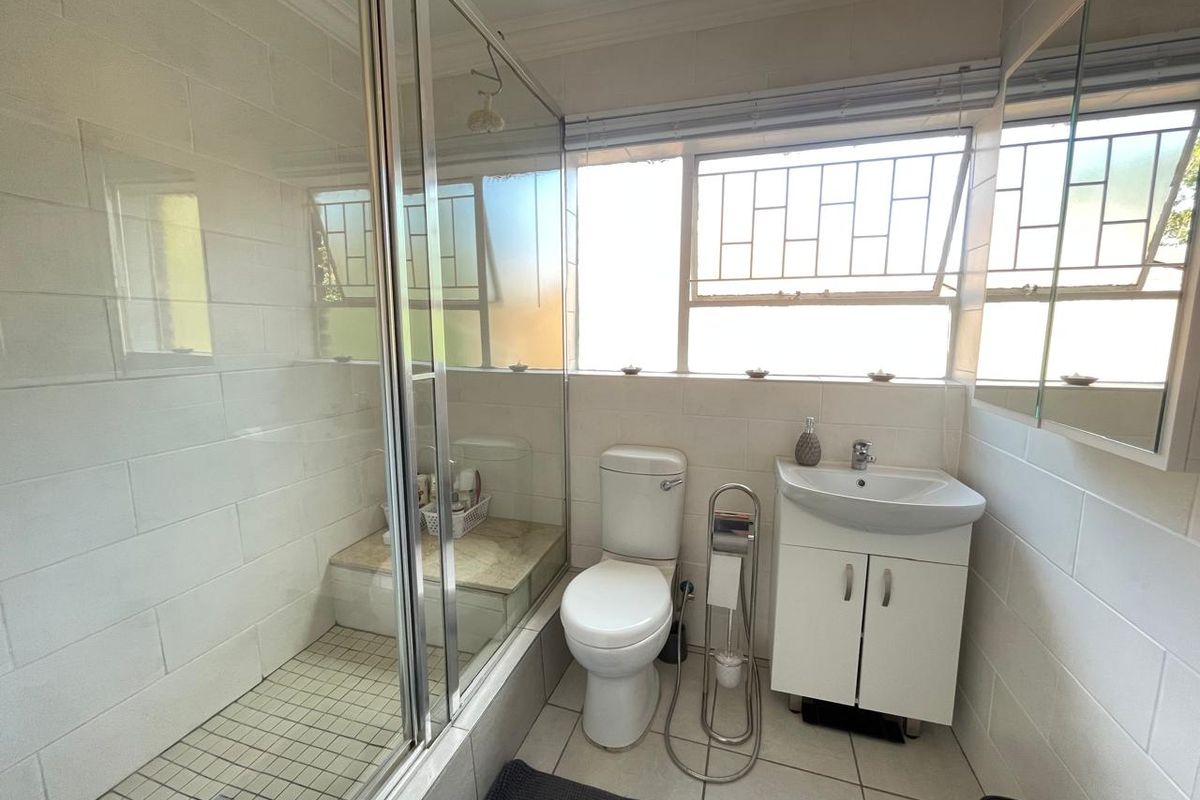 A functional bathroom featuring a glass-enclosed shower, a white vanity basin, and a standard toilet with security bars on the window.