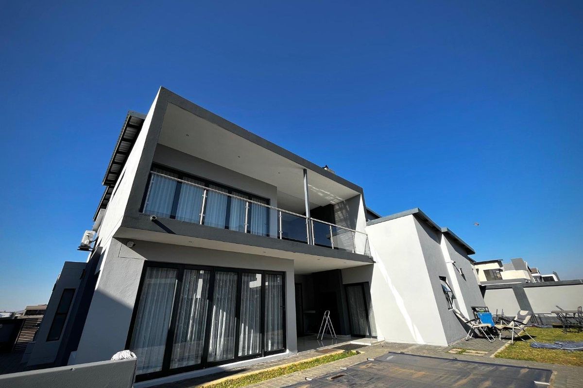 Rear view of a modern two-story house featuring a spacious balcony with glass balustrades, large sliding glass doors, and a paved patio area.