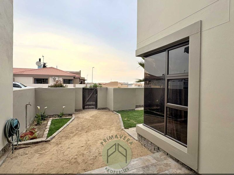 Rear exterior view of the property featuring a small garden area, boundary wall with a timber gate, and a tiled patio section.