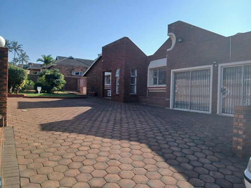Front view of a face brick house featuring a large paved driveway, sliding glass doors with security gates, and established garden surroundings.
